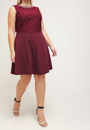 Femme marchant dans une robe sans manches bordeaux avec un corsage en dentelle et une jupe évasée, portant des talons à brides à la cheville beiges et noirs.