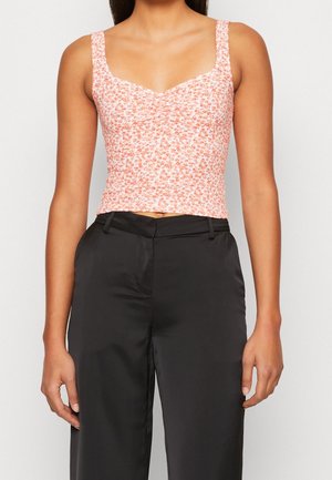 Kobieta ubrana w różowo-biały kwiecisty crop top bez rękawów, w połączeniu z czarnymi, szerokimi spodniami z wysokim stanem na jednolitym tle.