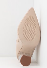 Beige pointed-toe shoe with textured sole and a low, transparent block heel resting against white walls.