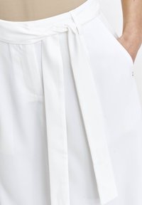 Biała spódnica z szerokim pasa, związana z przodu. Materiał wydaje się gładki z matowym wykończeniem. Zawiera boczne kieszenie dla funkcjonalności.