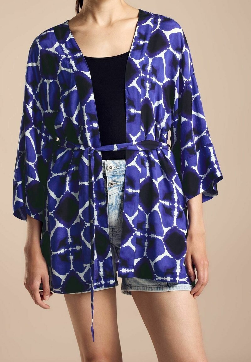 Blauwe en witte kimono met patroon, brede mouwen, een riem en een losse pasvorm, gedragen over een zwart top en denim shorts. Zachte stoftextuur.