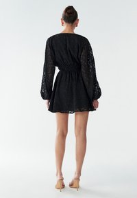 Juoda permatoma suknelė su faktūruotu raštu, ilgos pūstos rankovės, pabrėžta liemuo ir išplatėjusi apačia, derinama su smėlio spalvos aukštakulniais.