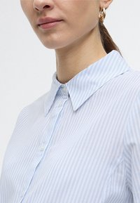 Lichtblauw gestreept overhemd met knopen, voorzien van een spitse kraag en zachte stof. Heeft een knoopsluiting aan de voorkant en opgerolde mouwen.