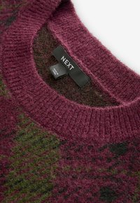 Gros plan sur l'encolure d'un pull en maille bordeaux et vert avec des étiquettes noires de la marque NEXT et Made in China.