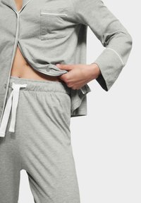 Grå bomulds pyjamas sæt består af en skjorte med knapper og en lomme samt tapered bukser med snor i taljen. Blød tekstur og kontrasterende hvide detaljer.