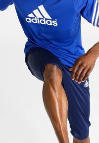 Person i blå Adidas sportkläder som lyfter ett ben, visar en muskulös vad och har handen vila nära låret.