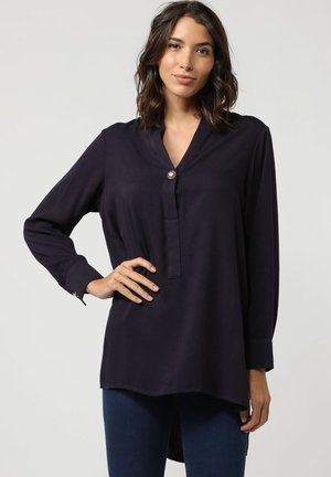 Blusa de manga larga de color navy con escote en V, detalle de botón único y puños enrollados; tela suave con un ajuste ligeramente holgado y un dobladillo trasero más largo.