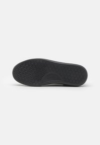 Semelle de chaussure noire avec motif de bande de roulement hexagonal, zone de grip texturée près de la boule du pied et léger maintien de la voûte plantaire visible.