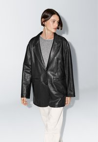 Černý kožený oversized blejzr se dvěma knoflíky, klopovým límečkem, bočními kapsami, nošený přes šedou košili, v kombinaci se světlými kalhotami.