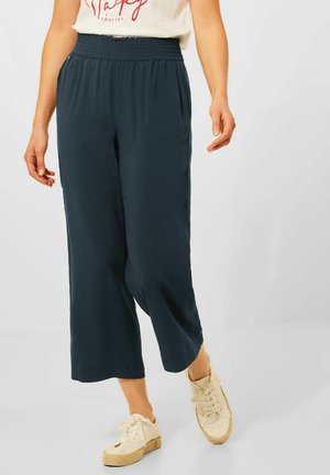 Vrouw die marineblauwe, wijd uitgesneden cropped broek draagt, witte sneakers en een wit T-shirt met rode tekst, staand op een lichte achtergrond.