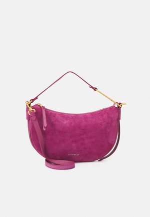 Compact roze suède handtas met gouden gespen, gebogen vorm, ritssluiting en twee verstelbare riemen om over de schouder of crossbody te dragen.