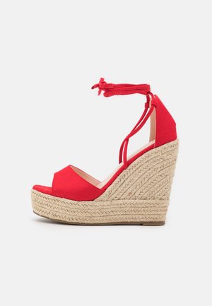 Sandale rouge en forme de coin avec bout ouvert, lanières à la cheville, et talon à plateforme en jute tressé sur un fond blanc uni.