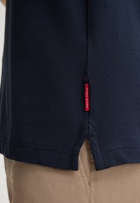 Marineblauwe polo met textuur, zijsplitsingen aan de zoom en een rode merklabel aan de linkerkant nabij de zoom.