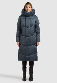 Tumehall polsterdatud pikk mantel kõrge kraega, millel on vertikaalsed õmblused ja kaks küljetaskut. Kandmisel mustad põlvest kõrgemad saapad.