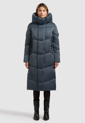 Abrigo largo acolchado en gris oscuro con cuello alto, con costuras verticales y dos bolsillos laterales. Se combina con botas negras hasta la rodilla.