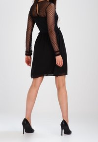 Černé šaty s průsvitnými dlouhými rukávy s puntíky a přiléhavým živůtkem, s jemnou splývavou sukní a vysokými podpatky se špičatou špičkou.