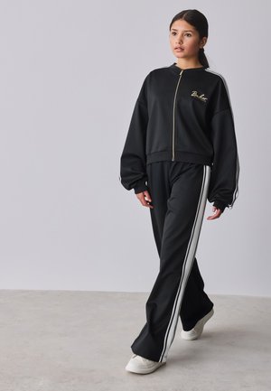 Junge Frau, die drinnen geht und eine schwarze Jacke mit Reißverschluss sowie passende Hose mit weißen Streifen und weiße Turnschuhe trägt.