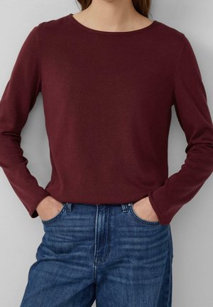 Personne portant une chemise à manches longues bordeaux et un jean bleu, les mains dans les poches, sur un fond gris uni.