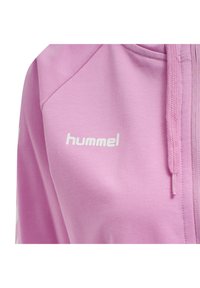 Ljusrosa zip-up hoodie med en slät textur; har en vit logotyp "hummel" och matchande dragsnöre på huvan.