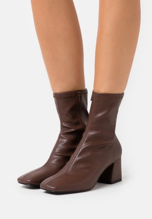 Brown leather ankle boots with block heels and side zippers worn on bare legs against a plain white background.