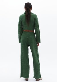 Zelený strukturovaný dvoudílný outfit, který zahrnuje zkrácený top s dlouhými rukávy a široké kalhoty. Měkká, žebrovaná látka s uvolněným střihem.