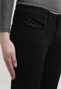 Jeans in denim neri con vestibilità slim, caratterizzati da un design a cinque tasche, cuciture a contrasto sottili e una texture liscia. Vista ravvicinata accanto a un maglione grigio a coste.