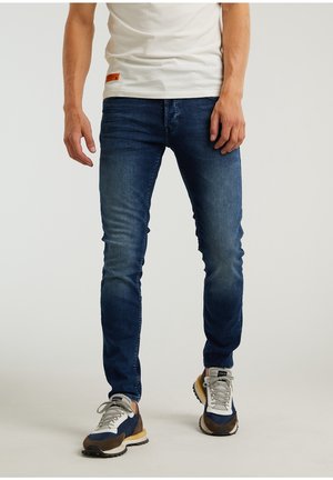 Mężczyzna noszący ciemnoniebieskie dopasowane dżinsy, białą koszulkę i niebiesko-biało-brązowe trampki, stojący na jasnym tle.