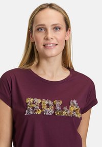 Burgunder T-Shirt mit dem Wort "HOLA", verziert mit strukturiertem Blumenapplikationen in Violett-, Gelb- und Grüntönen. Mit kurzen Ärmeln.
