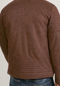 Braune strukturierte Jacke mit gerippten Details am unteren Rücken und an den oberen Ärmeln, getragen von einer Person in blauer Jeans, von hinten gezeigt.