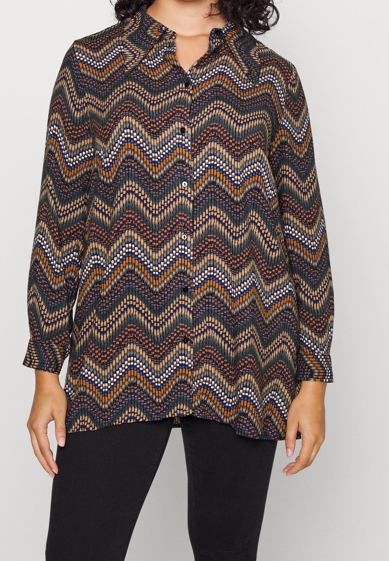 Femme portant une chemise à manches longues boutonnée avec un motif en zigzag multicolore en marron, orange, marine et blanc, associée à un pantalon noir.