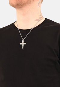 Sölv korset pendel på en kedja som bärs över en svart t-shirt. Korset har geometriska mönster och en polerad yta.