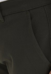 Dunkelgrüner, texturierter Stoff nahaufnahme von einer Hosentasche mit sichtbaren Nähdetails und einem schrägen Schnitt, der einen subtilen Glanz aufweist.