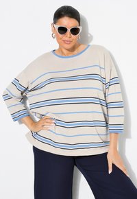 Beige raido yläpaita sinisten ja mustien vaakaraitojen kanssa, pyöreä kaula-aukko, tilava malli ja kolmanneshelma. Yhdistetty tummansinisiin housuihin.