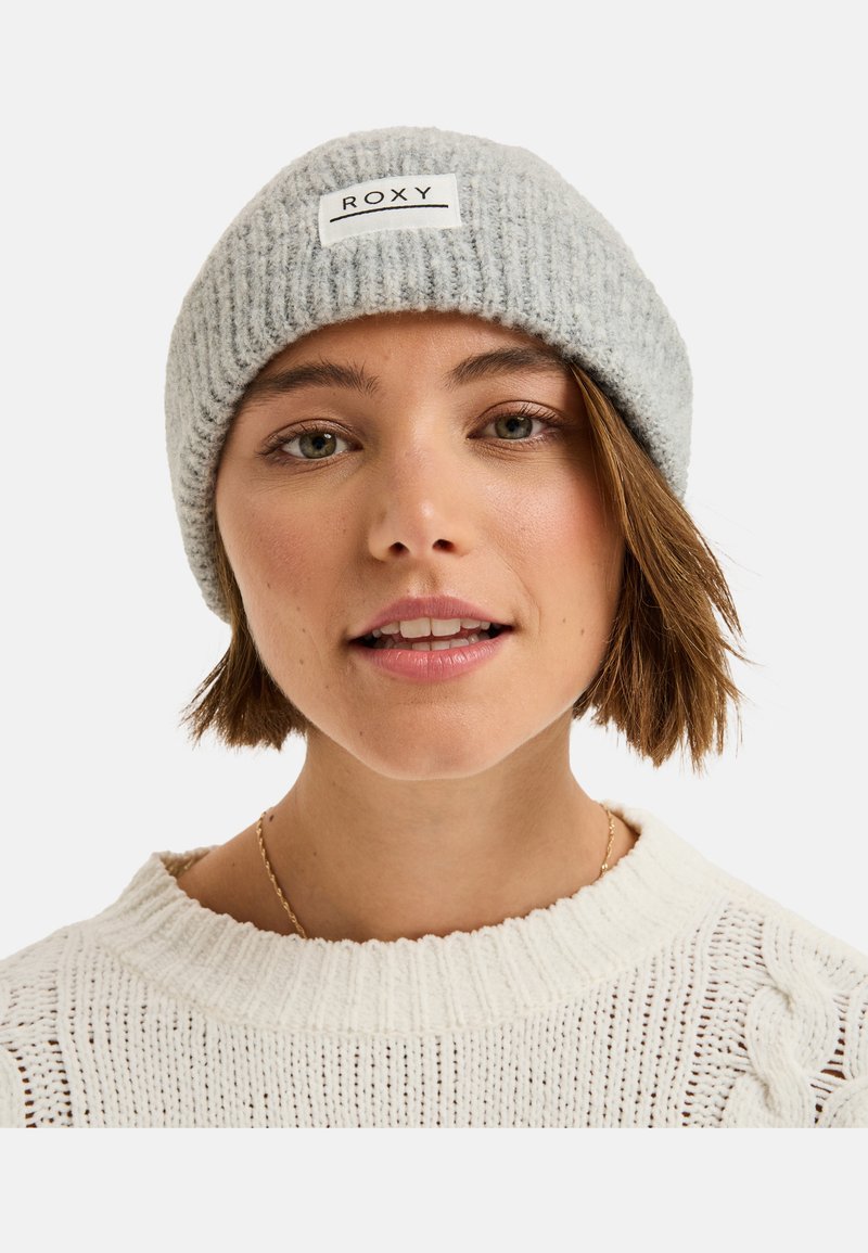 Grijze geribde gebreide beanie met een wit rechthoekig label met de tekst "ROXY." De stof lijkt zacht en gestructureerd, en past snugger op het hoofd.