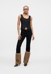 Γυναίκα με πλεξούδες στα μαλλιά και γυαλιά φοράει μαύρο αμάνικο τοπ με κουμπιά, μαύρο παντελόνι, καφέ ψεύτικα γούνινα καλύμματα ποδιών και μυτερά παπούτσια.