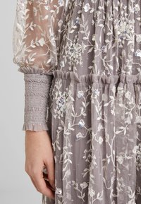 Robe en organza lavande avec broderie florale blanche et sequins, dotée de poignets froncés et d'une taille froncée, main posée à côté du corps.