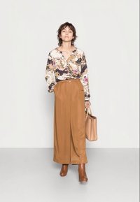 Blusa com padrão floral e mangas longas, combinada com calças largas de cintura alta castanhas e botas de tornozelo castanhas; segurando uma mala bege.