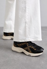 Baskets à imprimé léopard noir et beige avec un design en maille et cuir, dotées d'une semelle épaisse beige et de lacets, associées à un pantalon blanc.