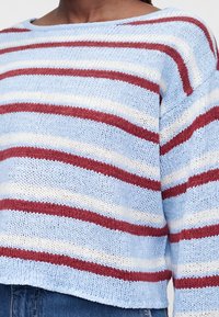 Personne portant un pull tricoté bleu clair avec des rayures blanches et rouges horizontales, associé à un jean bleu en denim.