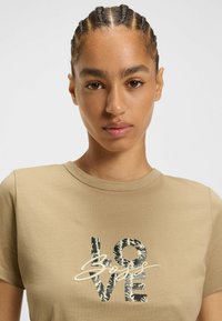 Beige t-shirt med korte ærmer, der prydes af et grafisk design af ordet "KÆRLIGHED" i sort, accentueret med en håndskrevet stil. Glat tekstur.