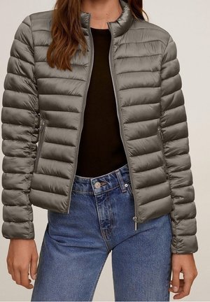 Femme portant une veste matelassée grise zippée sur un haut noir et un jean bleu, debout devant un fond neutre.