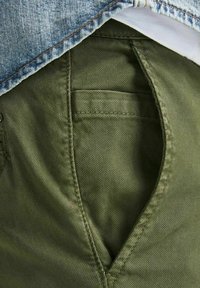 Olivgröna bomullbyxor med en framficka; texturerad tyg och synlig sömnad. Lager på med en denimjacka ovanpå.