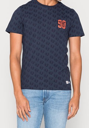 Mężczyzna w granatowej koszulce z geometrycznym wzorem i pomarańczowym nadrukiem "90" oraz w jasnoniebieskich dżinsach, stojący z rękami swobodnie opuszczonymi wzdłuż ciała.