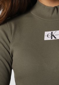 Oliivinvihreä ribbineulepaita, jossa on korkea kaula-aukko ja Calvin Klein Jeans -merkki näkyvästi etupuolella. Sileä rakenne ja muotoiltu istuvuus.