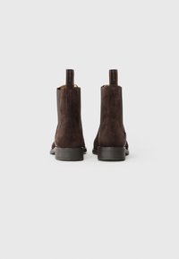 Bottines en daim marron avec une texture lisse, bout rond, talon bas empilé, et détail de tirette contrastante au niveau du talon.