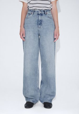 Person iført lyseblå højttaljede vidde jeans kombineret med sorte sko og en hvid stribet kortærmet skjorte.