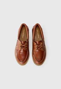 Brown leather shoes with a rounded toe, gold eyelets, and laces. The sole is beige with a stitched border, featuring a smooth texture.
