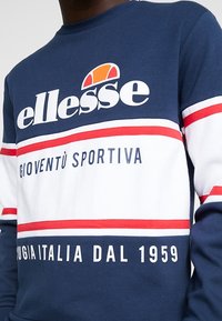 Marinblå och vit sweatshirt med djärva röda ränder. Har "ellesse"-logotyp, sportinspirerad text och en bekväm rundhalsdesign.
