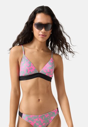 Femme aux longs cheveux bouclés portant de grandes lunettes de soleil foncées et un bikini rose et gris à motifs avec des bordures noires sur un fond uni.
