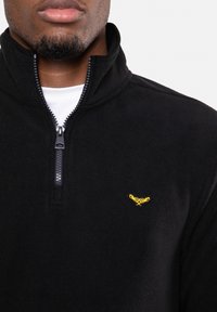 Pull-over en polaire noire avec un col haut, une fermeture à glissière partielle et un petit logo jaune brodé sur la poitrine gauche. Texture douce.